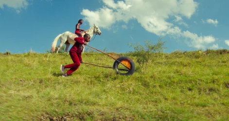 ANCESTRAL VISIONS OF THE FUTURE A child in red clothing rolls a burning tire down a green hill, while another child rides a white horse in the background.