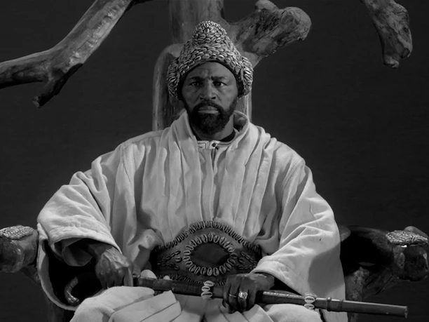 KATANGA, LA DANSE DES SCORPIONS A man in traditional dress sits seriously on an ornately carved chair.