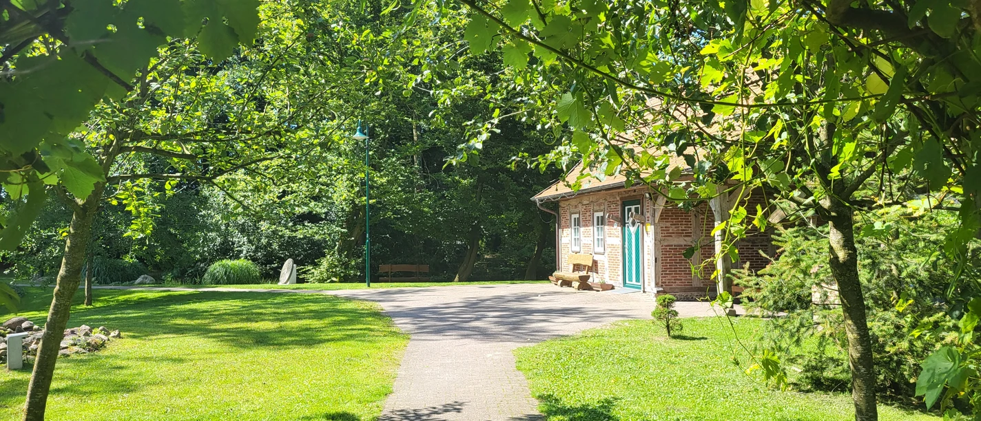 Insel Saßlage - Hümmling-Pfad Großer Sand, Holte-Lastrup ©Naturpark Hümmling.jpg Backsteinhaus mit grün-blauer Tür im Sonnenschein, umgeben von Bäumen und gepflegtem Rasen.