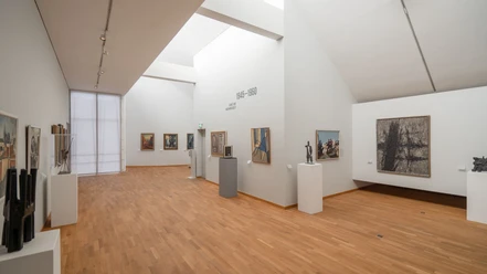 Innenraum eines Museums mit Bildern und Kunstwerken an den weißen Wänden und warmem Parkettboden.
