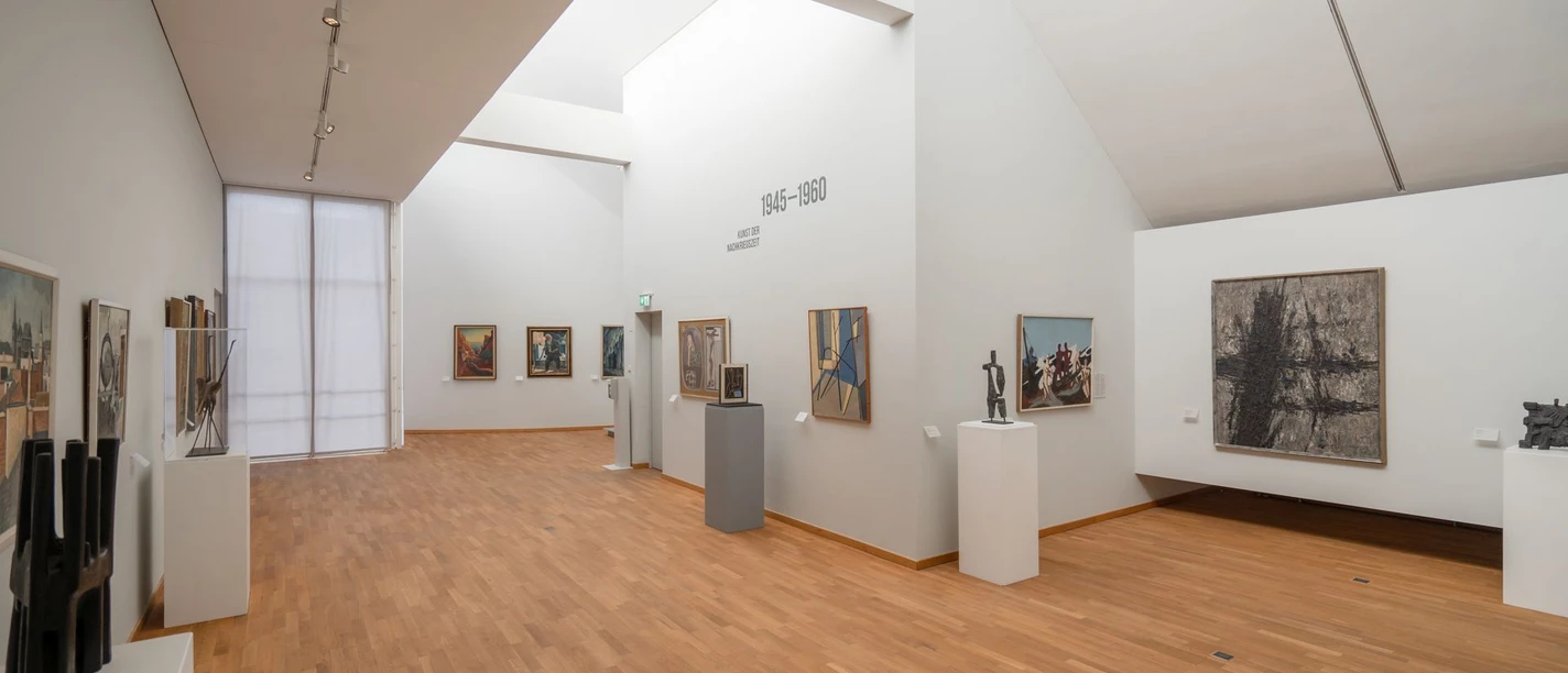 Innenraum eines Museums mit Bildern und Kunstwerken an den weißen Wänden und warmem Parkettboden.