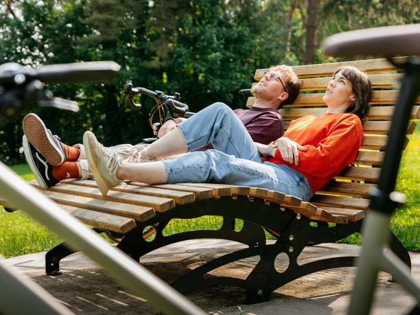 Pause an der Grenzgängerroute Twee mensen relaxen op een grote gebogen houten bank in een park.