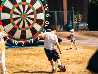 Even Flow Festival 2025 Eine Szene auf einer Wiese zeigt Kinder, die mit einem riesigen Dartboard aus Stoff spielen. Ein Junge tritt stolz gegen den Ball, während andere zuschauen. Die Farben sind lebendig und die Atmosphäre fröhlich und unbeschwert.A scene on a meadow shows children playing with a giant fabric dartboard. A boy proudly kicks the ball while others look on. The colors are vibrant and the atmosphere is cheerful and carefree.
