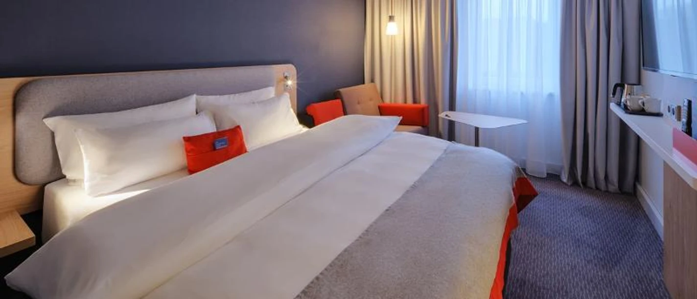 Holiday Inn Express Frankfurt Messe Ein modernes Hotelzimmer mit großem Bett, dunkler Wand und roten Akzenten.