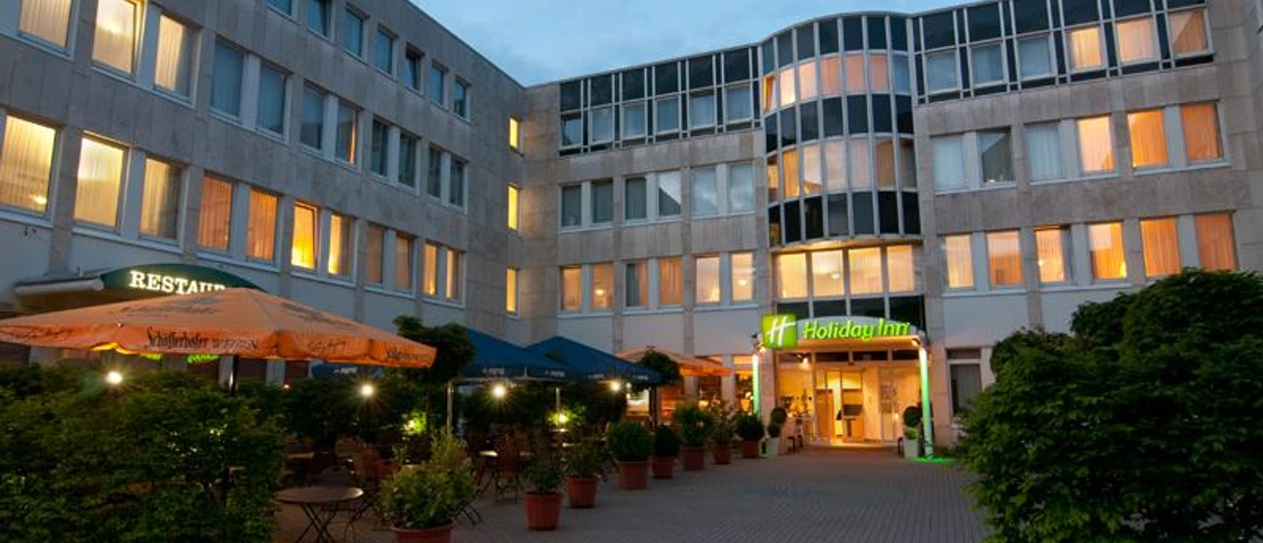 Holiday Inn Frankfurt Airport-Neu-Isenburg Ein Holiday Inn Hotel bei Dämmerung mit beleuchteten Fenstern und Terrasse.