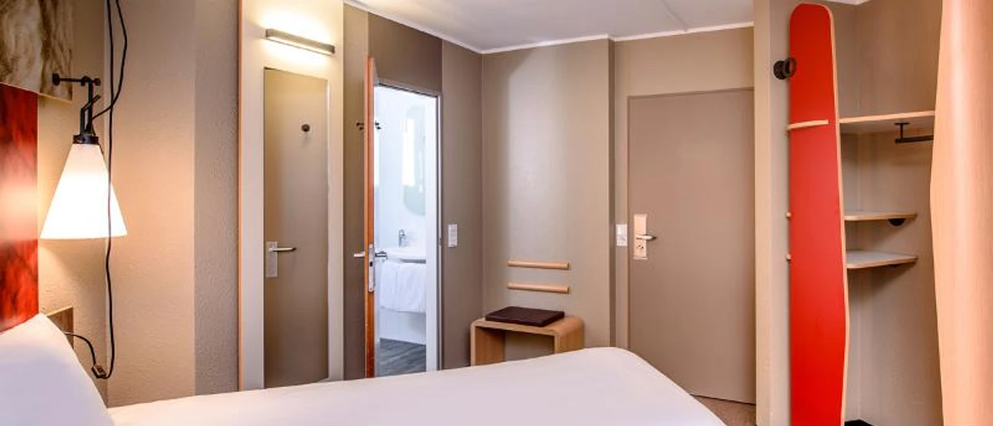 Ibis Frankfurt Centrum Ein kleines Hotelzimmer mit zwei Betten, rotem Dekor und Badezimmerzugang.