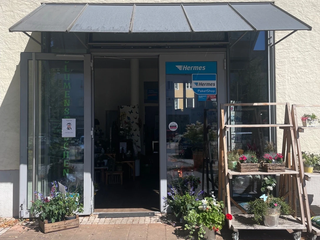 blumenstuebchen.jpg Das Bild zeigt den Eingang eines Blumenladens mit offenen Glastüren, umrahmt von blühenden Pflanzen.