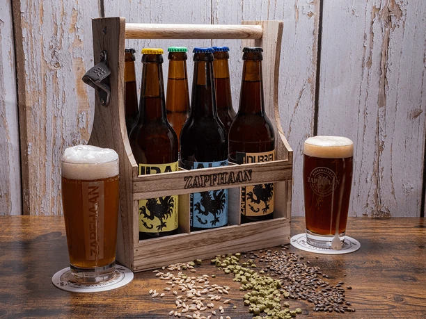 Rustikale Sixpackträger aus Holz von Zapfhaan aus Haan Sechs bunte Bierflaschen in einem hölzernen Sixpackträger mit Zapfhaan-Schriftzug, umgeben von Hopfen und Malz.