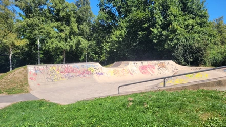 Die Skaterbahn in Lübbecke ist umgeben von üppigen Bäumen und bunter Graffiti-Kunst. Farbige Graffitis zieren die Rampen und Wände, während das grüne Gras und der strahlend blaue Himmel eine angenehme Kulisse bilden.