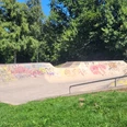 Die Skaterbahn in Lübbecke ist umgeben von üppigen Bäumen und bunter Graffiti-Kunst. Farbige Graffitis zieren die Rampen und Wände, während das grüne Gras und der strahlend blaue Himmel eine angenehme Kulisse bilden.