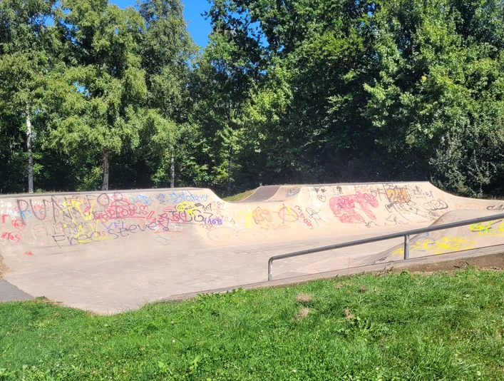 Skateranlage an der Kreissporthalle Lübbecke Die Skaterbahn in Lübbecke ist umgeben von üppigen Bäumen und bunter Graffiti-Kunst. Farbige Graffitis zieren die Rampen und Wände, während das grüne Gras und der strahlend blaue Himmel eine angenehme Kulisse bilden.