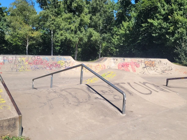 Skateranlage an der Kreissporthalle Lübbecke Die Skaterbahn in Lübbecke ist umgeben von üppigen Bäumen und bunter Graffiti-Kunst. Farbige Graffitis zieren die Rampen und Wände, während das grüne Gras und der strahlend blaue Himmel eine angenehme Kulisse bilden.