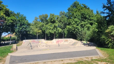Skateranlage an der Kreissporthalle Lübbecke Die Skaterbahn in Lübbecke ist umgeben von üppigen Bäumen und bunter Graffiti-Kunst. Farbige Graffitis zieren die Rampen und Wände, während das grüne Gras und der strahlend blaue Himmel eine angenehme Kulisse bilden.