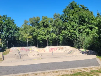 Skateranlage an der Kreissporthalle Lübbecke Die Skaterbahn in Lübbecke ist umgeben von üppigen Bäumen und bunter Graffiti-Kunst. Farbige Graffitis zieren die Rampen und Wände, während das grüne Gras und der strahlend blaue Himmel eine angenehme Kulisse bilden.
