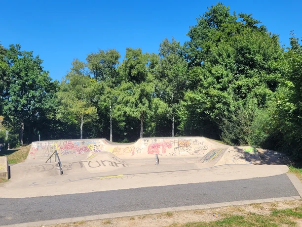 Skateranlage an der Kreissporthalle Lübbecke Die Skaterbahn in Lübbecke ist umgeben von üppigen Bäumen und bunter Graffiti-Kunst. Farbige Graffitis zieren die Rampen und Wände, während das grüne Gras und der strahlend blaue Himmel eine angenehme Kulisse bilden.