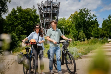 Am Baumwipfelpfad in Bad Iburg Twee mensen op e-bikes voor een uitkijktoren op het platteland op een zonnige dag.
