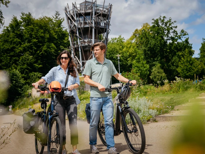 Am Baumwipfelpfad in Bad Iburg Zwei Menschen fahren bei sonnigem Wetter mit E-Bikes vor einem Aussichtsturm im Grünen.Two people riding e-bikes in front of a lookout tower in the countryside on a sunny day.To personer på elcykler foran et udsigtstårn på landet på en solrig dag.Twee mensen op e-bikes voor een uitkijktoren op het platteland op een zonnige dag.