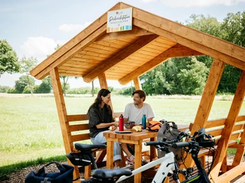 Picknickpause an der Friedensroute Ein überdachter Holzpavillon auf einer Wiese mit zwei Personen und Fahrrädern im Vordergrund.A roofed wooden pavilion on a meadow with two people and bicycles in the foreground.En overdækket træpavillon på en eng med to personer og cykler i forgrunden.Een overdekt houten paviljoen op een weiland met twee mensen en fietsen op de voorgrond.