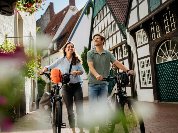 Unterwegs in der Altstadt von Osnabrück To personer går med cykler gennem en brostensbelagt gade med bindingsværkshuse.