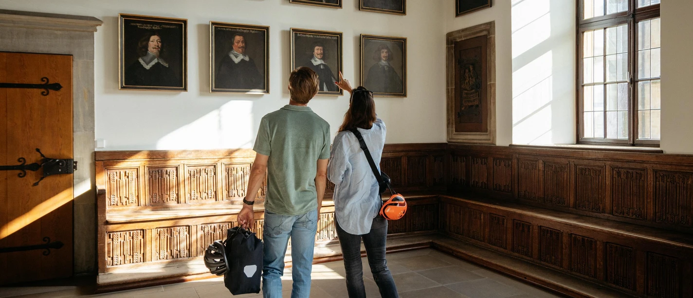 Der Friedenssaal im Osnabrücker Rathaus Two visitors look at paintings in a historic room with wood paneling and large windows.