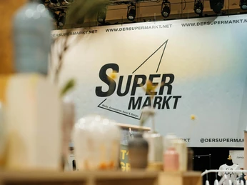 Der Super Weihnachtsmarkt Moderner und stilvoller Stand auf einem Weihnachtsmarkt. Im Vordergrund stehen stilvolle Glasgefäße auf einem Regal, während im Hintergrund eine große Werbewand mit dem Schriftzug "Super Markt" zu erkennen ist.Modern and stylish stand at a Christmas market. In the foreground are stylish glass jars on a shelf, while a large advertising wall with the words "Super Markt" can be seen in the background.