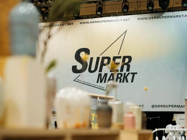 Modern and stylish stand at a Christmas market. In the foreground are stylish glass jars on a shelf, while a large advertising wall with the words "Super Markt" can be seen in the background.
