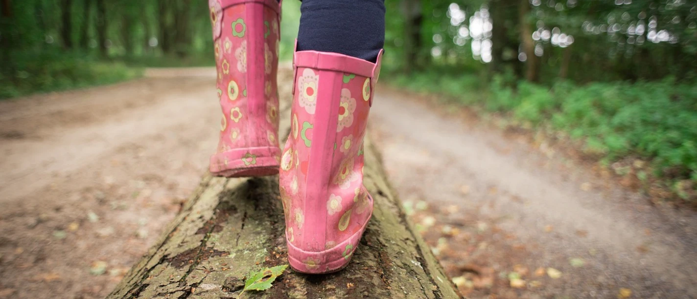 Rosafarbene Gummistiefel auf einem Baumstamm in einem Wald. Person balanciert entlang des Stammes.
