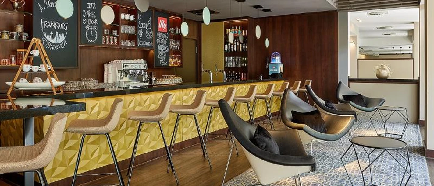 Le Meridien Frankfurt Eine moderne Hotelbar mit gelber Theke, Barhockern und Sitzgelegenheiten.