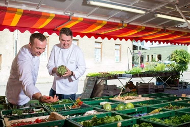 koeche-bauernmarkt-bad-aibling-einkaufen.jpg Zwei Köche wählen frisches Gemüse am Bauernmarkt Bad Aibling aus; farbenfrohe Marktatmosphäre.