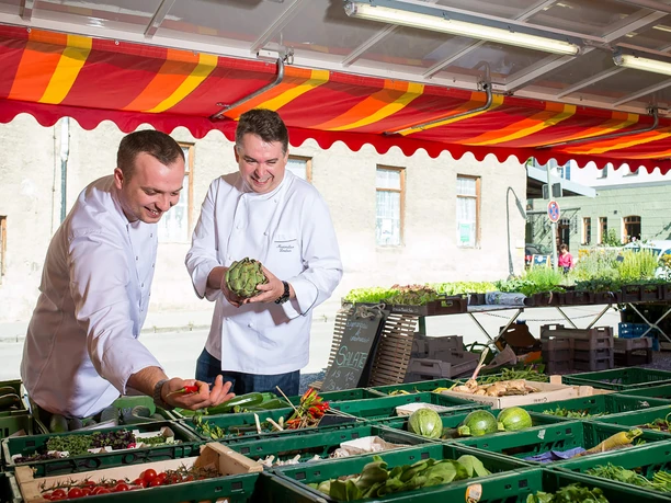 koeche-bauernmarkt-bad-aibling-einkaufen.jpg Zwei Köche wählen frisches Gemüse am Bauernmarkt Bad Aibling aus; farbenfrohe Marktatmosphäre.