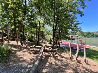 Niedrigseilgarten in Heiligenhaus Niedrigseilgarten aus Seilen und Holzstämmen unter schattigen Bäumen, neben einem Sportplatz gelegen.