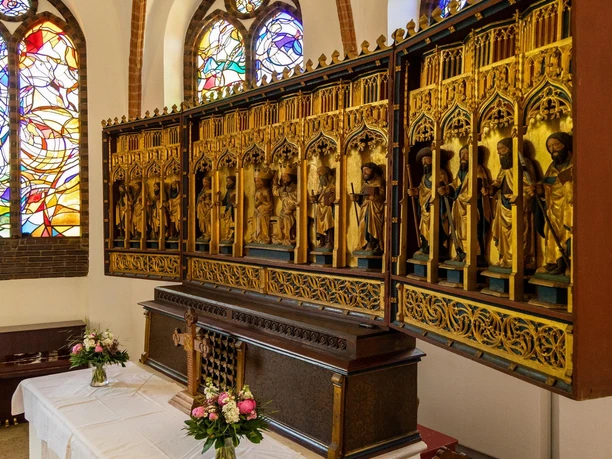 Kirche In einer Kirche steht ein Holzaltar mit geschnitzten Figuren. Buntglasfenster im Hintergrund werfen farbige Lichtmuster. Auf dem Altar befinden sich Blumen und ein Holzkreuz.