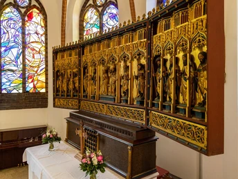 Amelinghausen_Betzendorf_st_Peter_Paul_Kirche_ccbysa_tl_20230531-7.JPG In einer Kirche steht ein Holzaltar mit geschnitzten Figuren. Buntglasfenster im Hintergrund werfen farbige Lichtmuster. Auf dem Altar befinden sich Blumen und ein Holzkreuz.