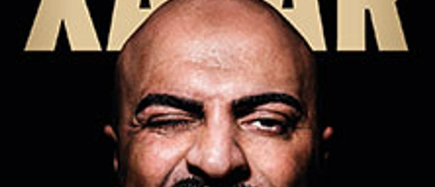 Das Bild zeigt eine frontal aufgenommene Nahaufnahme des muskulären Gesichts eines Mannes mit Glatze, der eine Grimasse zieht und die Zähne zeigt.