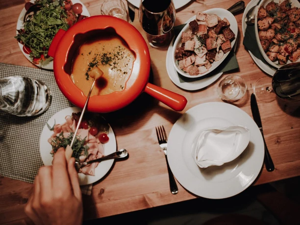 Uriger-Baudenabend Ein rustikales Fondue auf Holztisch, umgeben von Beilagen, Kräutern und Rotwein in gemütlicher Atmosphäre.