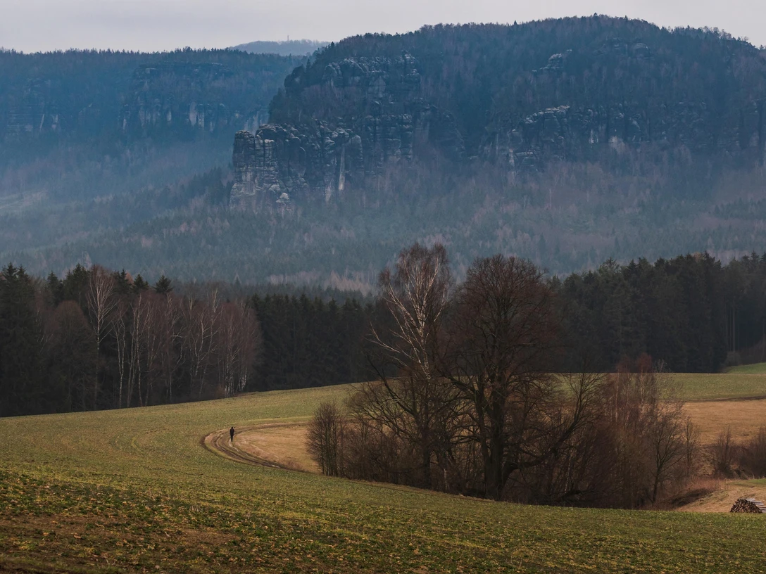 Panoramaweg Wanderweg durch Felder mit Blick auf bewaldete Felsformation im Hintergrund bei bewölktem Himmel.Hiking trail through fields with a view of a wooded rock formation in the background under a cloudy sky.Turistická stezka přes pole s výhledem na zalesněný skalní útvar v pozadí pod zataženou oblohou.Szlak pieszy przez pola z widokiem na zalesioną formację skalną w tle pod zachmurzonym niebem.Wandelpad door velden met uitzicht op een beboste rotsformatie op de achtergrond onder een bewolkte hemel.Sentiero escursionistico attraverso i campi con vista su una formazione rocciosa boscosa sullo sfondo, sotto un cielo nuvoloso.