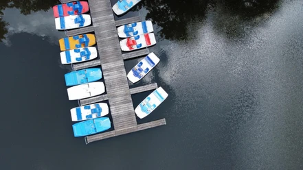 Lopausee2025 Mehrere bunte Boote liegen vertäut an einem Holzsteg auf einem ruhigen See. Die Wasseroberfläche spiegelt die Wolken wider. Die Boote sind in Blau, Rot und Gelb bemalt.