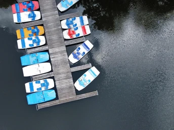 50-jahre-samtgemeinde-amelinghausen-cover (40).JPG Mehrere bunte Boote liegen vertäut an einem Holzsteg auf einem ruhigen See. Die Wasseroberfläche spiegelt die Wolken wider. Die Boote sind in Blau, Rot und Gelb bemalt.