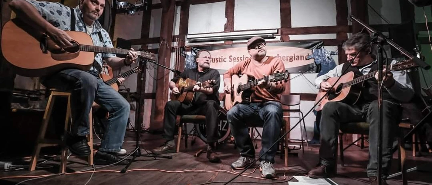 C81B25D6-DB30-42EC-9569-61DF647AC034.jpeg Männer mit Gitarren im Halbkreis sitzend vor einer Fachwerkwand