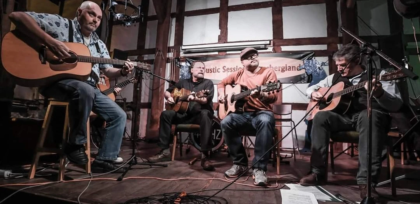 C81B25D6-DB30-42EC-9569-61DF647AC034.jpeg Männer mit Gitarren im Halbkreis sitzend vor einer Fachwerkwand