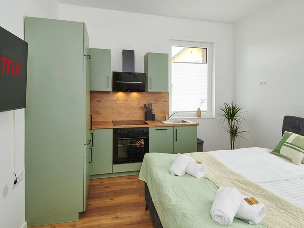 Wohn-/Schlafbereich mit Küchenzeile Wohn-/Schlafbereich mit grüner Küchenzeile, Doppelbett, TV, Fenster mit Tageslicht und Handtüchern.