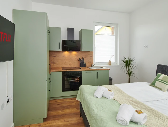 Wohn-/Schlafbereich mit Küchenzeile Wohn-/Schlafbereich mit grüner Küchenzeile, Doppelbett, TV, Fenster mit Tageslicht und Handtüchern.