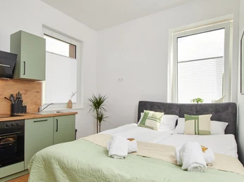 Schlafbereich Heller Raum mit Doppelbett, grüner Küche und großen Fenstern. Elegante Einrichtung und moderne Geräte.