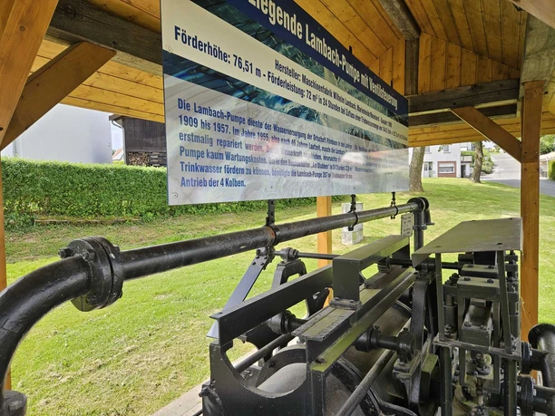 Historische Lambach-Pumpe in Pömbsen Historische Lambach-Pumpe aus schwarzem Metall mit großem Infoschild unter Holzdach.