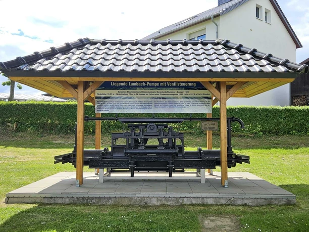 Lambach-Pumpe in Pömbsen Die historische Lambach-Pumpe steht unter einem hölzernen Dach auf einer steinernen Plattform.