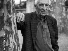 Ein älterer Mann lehnt in einem Park an einem Baum, blickt ernst und würdevoll in die Kamera.