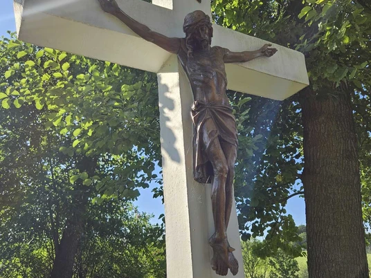 Kreuz nahe des Heiligenteiches in Pömbsen Kruzifix aus Holz mit Sonnenstrahlen, umgeben von grünem Laub.