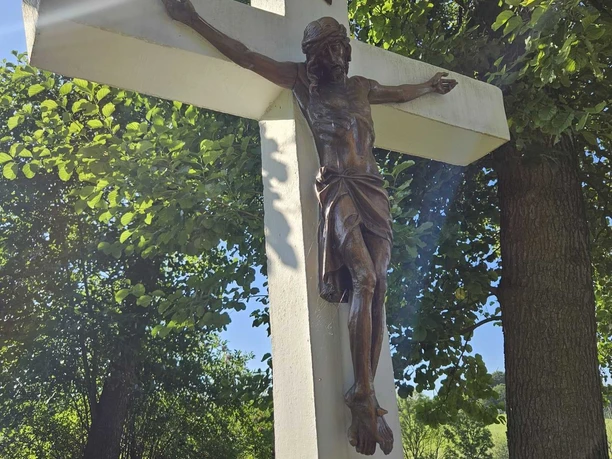 Kreuz nahe des Heiligenteiches in Pömbsen Kruzifix aus Holz mit Sonnenstrahlen, umgeben von grünem Laub.