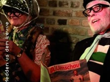 Zwei Personen in traditioneller rheinischer Verkleidung mit Brille und Kopftuch vor einer Backsteinwand.