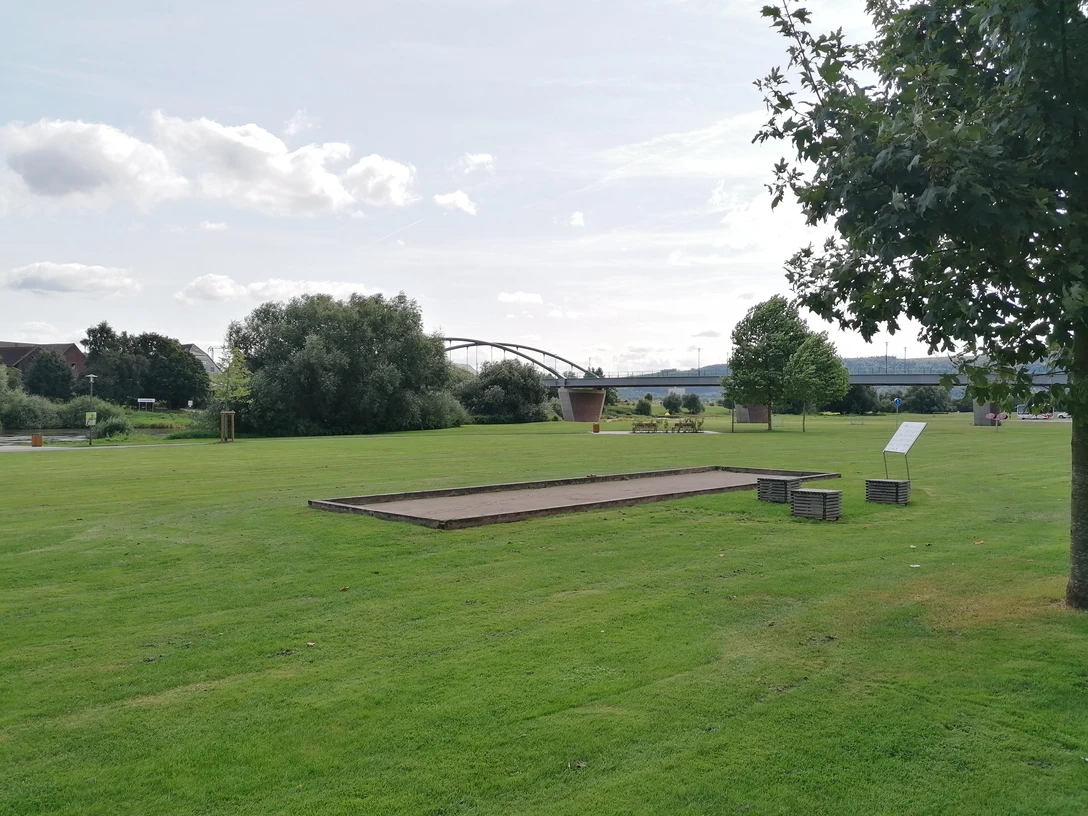 Boulebahn Beverungen Eine Boulebahn auf einer großen Rasenfläche mit Sitzwürfeln und einer Informationstafel daneben, im Hintergrund die Weser und die Weserbrücke.